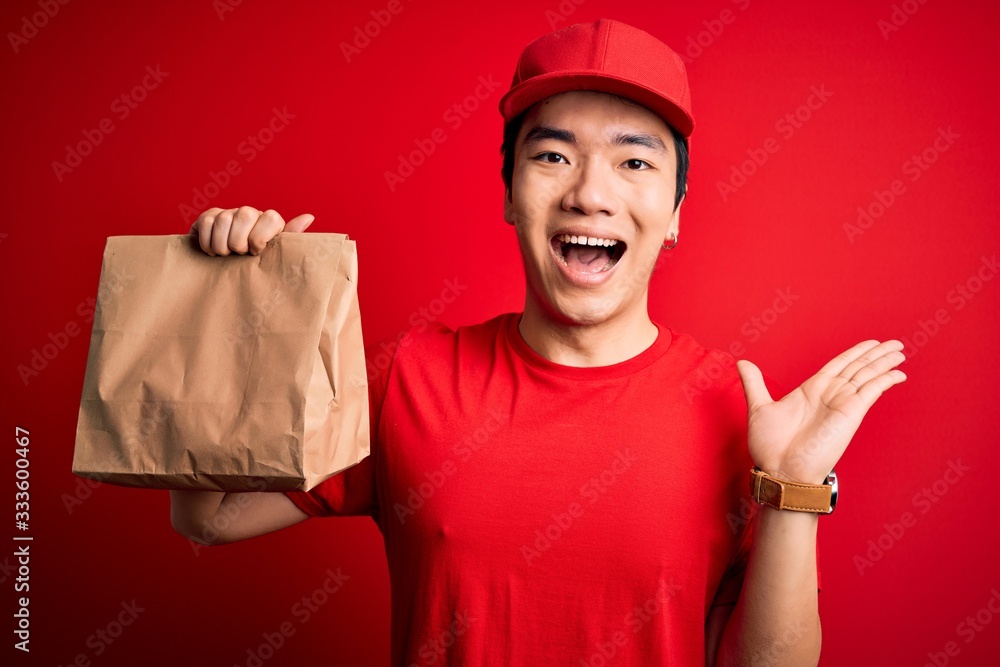 Young handsome chinese delivery man holding takeaway paper bag with food very happy and excited, winner expression celebrating victory screaming with big smile and raised hands Young handsome chinese delivery man holding takeaway paper bag with food very happy and excited, winner expression celebrating victory screaming with big smile and raised hands