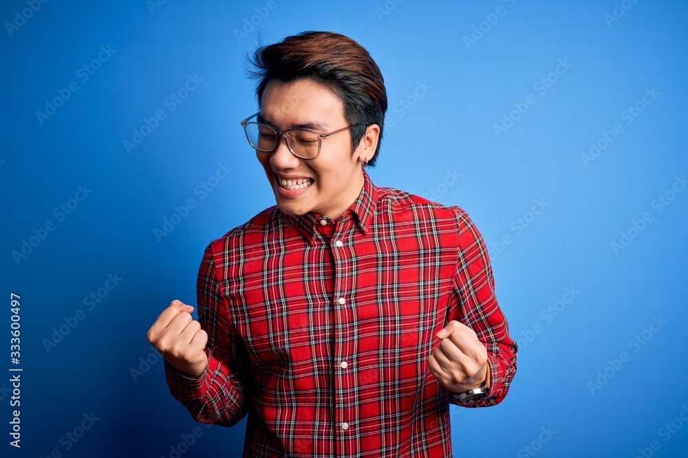Naklejka premium Young handsome chinese man wearing casual shirt and glasses over blue background very happy and excited doing winner gesture with arms raised, smiling and screaming for success. Celebration concept.