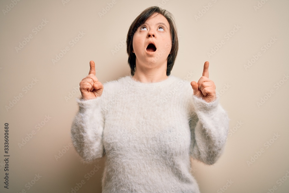 Young down syndrome woman standing over isolated background amazed and surprised looking up and pointing with fingers and raised arms. Young down syndrome woman standing over isolated background amazed and surprised looking up and pointing with fingers and raised arms.
