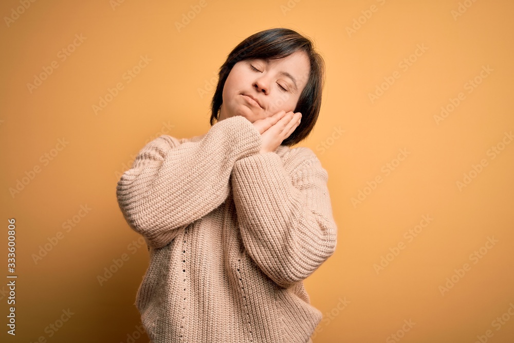 Young down syndrome woman wearing casual sweater over yellow background sleeping tired dreaming and posing with hands together while smiling with closed eyes. Young down syndrome woman wearing casual sweater over yellow background sleeping tired dreaming and posing with hands together while smiling with closed eyes.