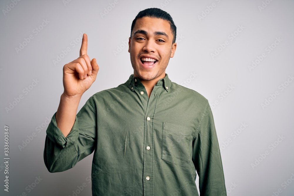 Young brazilian man wearing casual shirt standing over isolated white background pointing finger up with successful idea. Exited and happy. Number one. Young brazilian man wearing casual shirt standing over isolated white background pointing finger up with successful idea. Exited and happy. Number one.