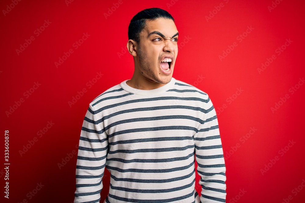 Young brazilian man wearing casual striped t-shirt standing over isolated red background angry and mad screaming frustrated and furious, shouting with anger. Rage and aggressive concept. Young brazilian man wearing casual striped t-shirt standing over isolated red background angry and mad screaming frustrated and furious, shouting with anger. Rage and aggressive concept.