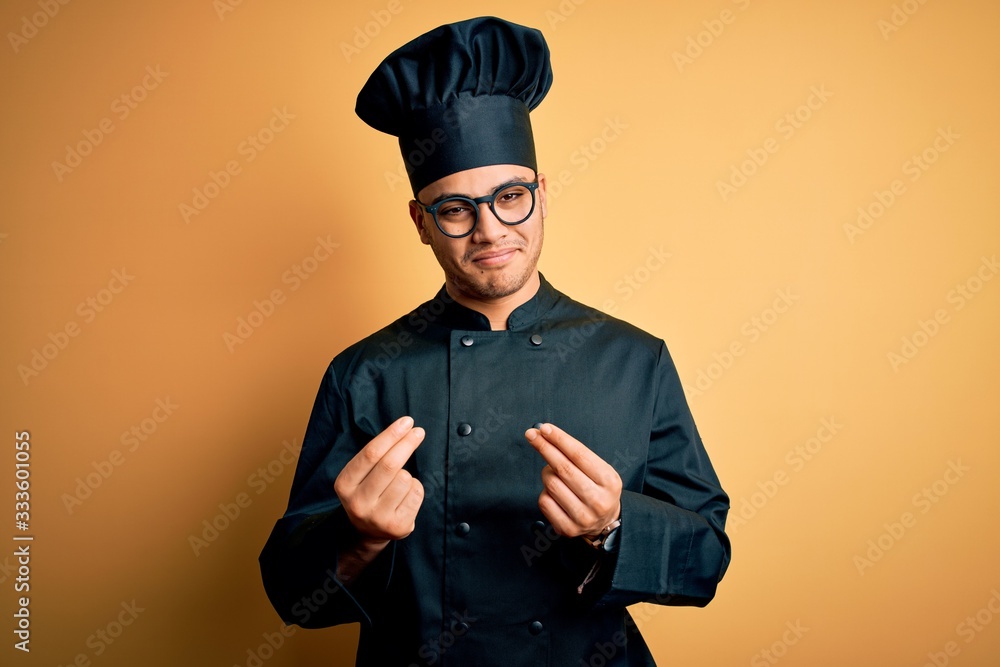 Young brazilian chef man wearing cooker uniform and hat over isolated yellow background doing money gesture with hands, asking for salary payment, millionaire business Young brazilian chef man wearing cooker uniform and hat over isolated yellow background doing money gesture with hands, asking for salary payment, millionaire business