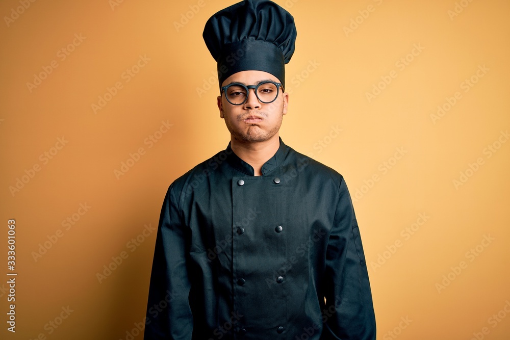 Young brazilian chef man wearing cooker uniform and hat over isolated yellow background puffing cheeks with funny face. Mouth inflated with air, crazy expression. Young brazilian chef man wearing cooker uniform and hat over isolated yellow background puffing cheeks with funny face. Mouth inflated with air, crazy expression.