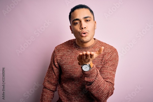 Young brazilian man wearing casual sweater standing over isolated pink background looking at the camera blowing a kiss with hand on air being lovely and sexy. Love expression.