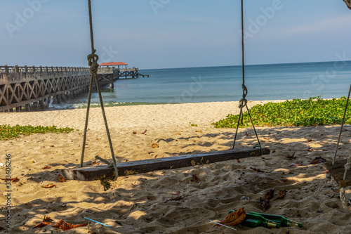 A simple wooden swing is hanging next to a clean tropical beach with blue sky background