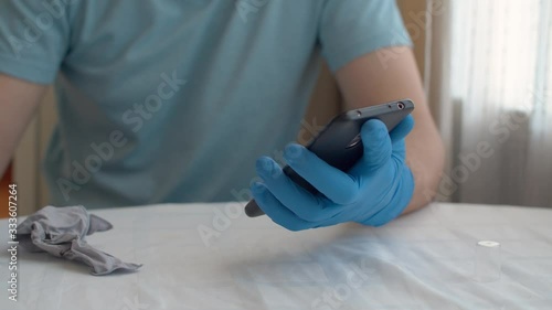 Close up male hands in protective gloves sanitaizing his smart phone. She is spraying sanitizer on it and wipes with a cloth. Hygiene during coronavirus epidemic. Concept of safety life. photo