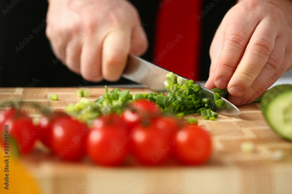 Cook holds knife in hand and cuts on cutting board green onions for salad or fresh vegetable soup with vitamins. Raw food and vegetarian recipe book in modern society popular concept. Cook holds knife in hand and cuts on cutting board green onions for salad or fresh vegetable soup with vitamins. Raw food and vegetarian recipe book in modern society popular concept.