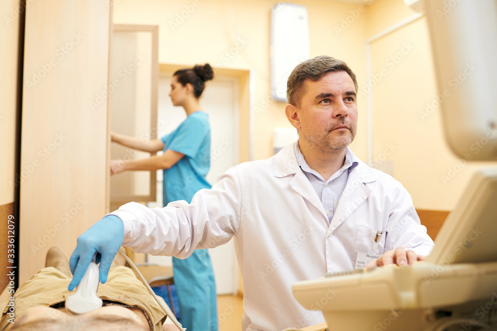 Fototapeta premium Serious mature doctor in lab coat doing ultrasound scanning while examining abdomen of patient