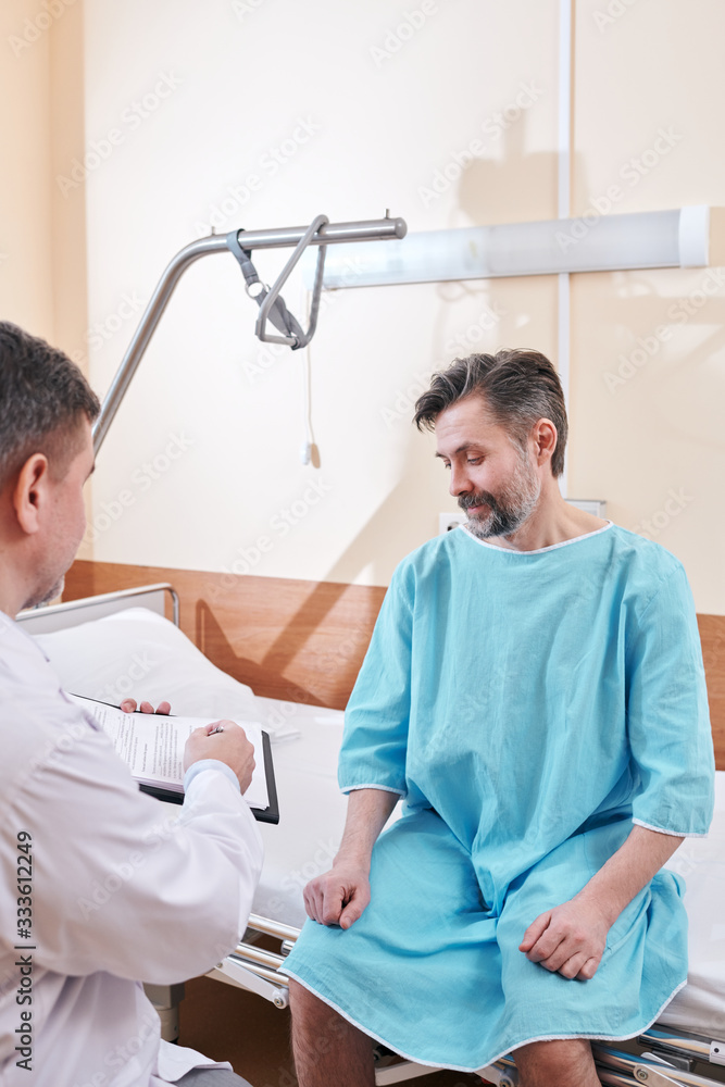 Obraz premium Over shoulder view of doctor pointing at place for signature while asking patient in hospital gown to sign medical agreement before operation