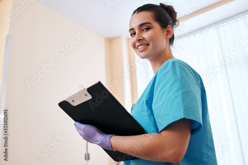 Portrait of smiling attractive nurse in surgical gloves making notes in medical card while doing rounds in hospital