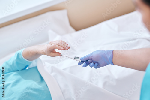 Above view of unrecognizable nurse in surgical gloves giving digital thermometer to patient in bed © pressmaster