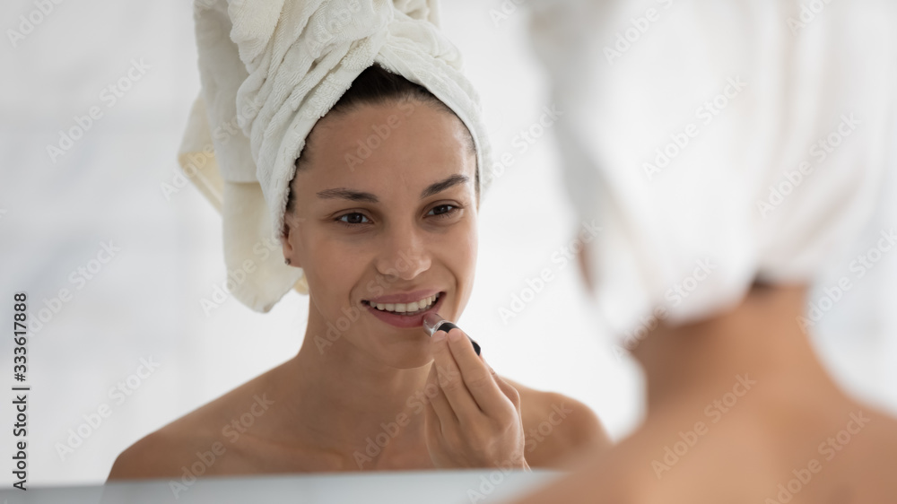 After morning shower head wrapped in towel attractive 35s woman looking in mirror holding lipstick tube applying long-lasting pink color on lips in bathroom. Concept of beauty and make up cosmetics ad