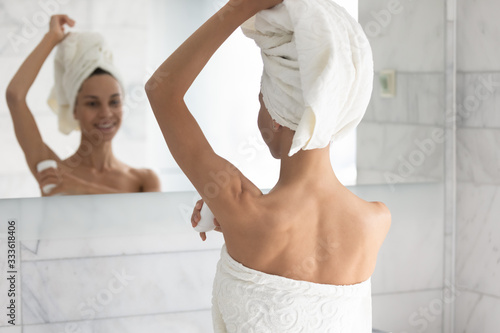 Rear view woman after shower body and head wrapped in towels reflected in mirror, apply at armpit anti perspirant stick, protect sweating cosmetic product, personal hygiene, grooming self-care concept photo