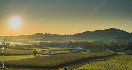 Beautiful morning panorama of a village or city of Sankt Georgen in Attergau, Austria. Visble sun with rays rising above the houses and green meadows.