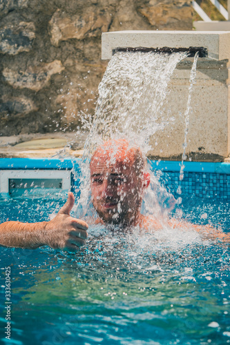 Young caucasian male with bald head enjoying a flow of water fountain flowing onto his head inside a swimming pool. Man showing thumbs up to a camera photo