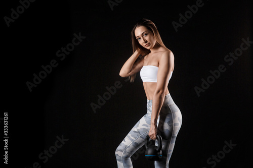 Fitness exercise woman holding kettlebell strength training biceps. Beautiful sweaty fitness instructor looking. Mixed race Caucasian female sweating isolated on black background