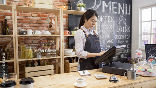 smiling shop assistant using pos point of sale terminal to put in order from note at restaurant register. young girl waitress standing in wooden bar counter and working on digital tablet in cafe