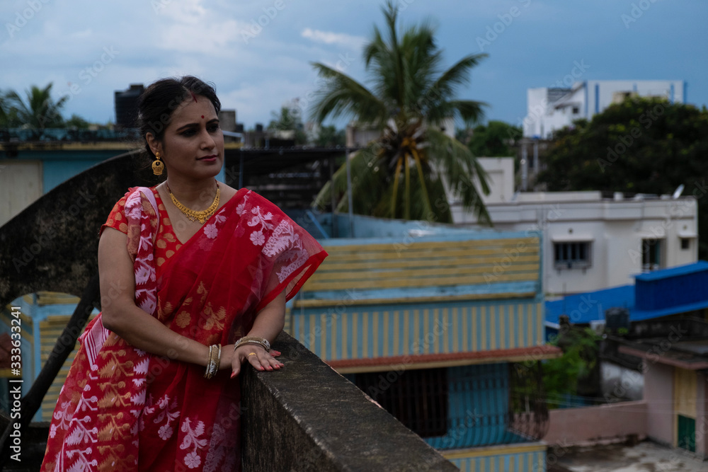 Portrait of a beautiful smiling brunette Indian Bengali woman in traditional red sari standing on the roof top under blue sky in afternoon of Durga Puja festival in urban background. Indian lifestyle