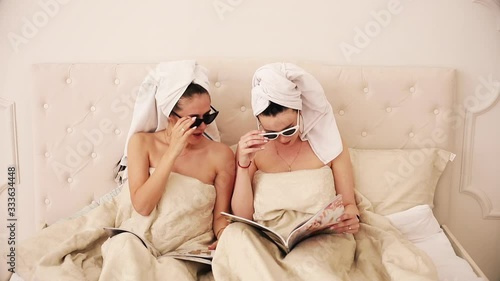 Portrait of thwo friends with white towels on their heads and with sunglasses lie on a bed under the white blanket, read magazines and surprised by something