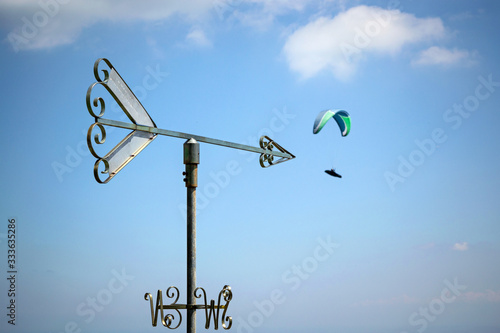 Arrow shape weathervane against summer's sky with cumulus and paragliders in background, italian Alps. Italy