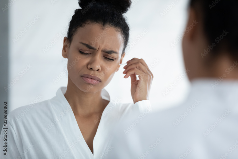 Unhealthy young African woman hold cotton bud cleaning ears removes earwax feels discomfort and pain. Earache caused by incorrect bad hygiene, infection, otitis media symptoms, health problems concept Unhealthy young African woman hold cotton bud cleaning ears removes earwax feels discomfort and pain. Earache caused by incorrect bad hygiene, infection, otitis media symptoms, health problems concept