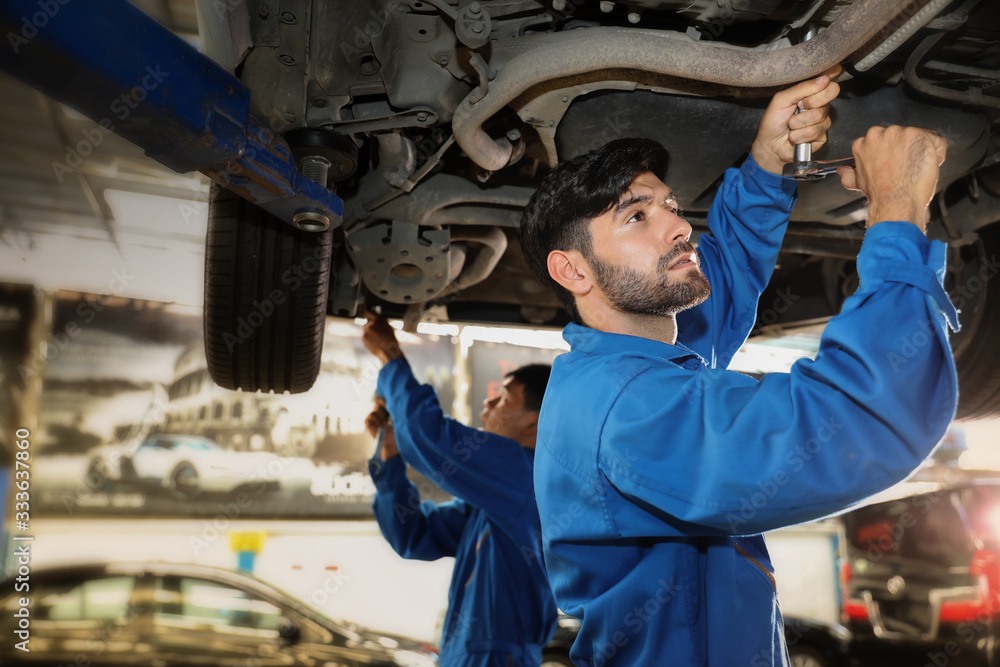Mechanic using screwdriver for repair with repair equipment standing under the car in maintainance service center which is a part of showroom.Car maintenance and repair concept. Mechanic using screwdriver for repair with repair equipment standing under the car in maintainance service center which is a part of showroom.Car maintenance and repair concept.