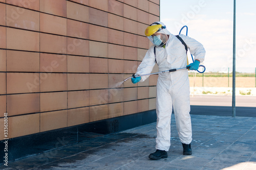 Man wearing an NBC personal protective equipment (ppe) suit, gloves, mask, and face shield, cleaning the streets with a backpack of pressurized spray disinfectant water to remove covid-19 coronavirus.