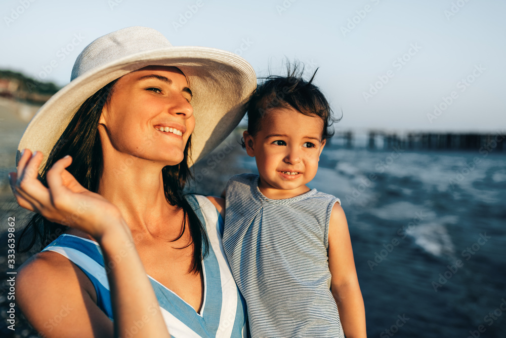 Smiling beautiful mother with cute daughter wearing white hat and striped dress posing at the sea. Happy female with little girl looking to the ocean. Caucasian female with baby posing at ocean sunset