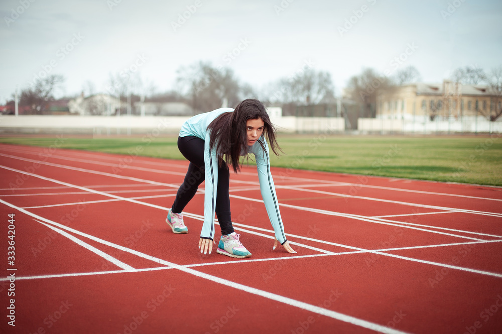 A beautiful girl performs sports exercises in a blue stadium with brown rubber tracks. Woman in a blue trowel and black leggings. A beautiful girl performs sports exercises in a blue stadium with brown rubber tracks. Woman in a blue trowel and black leggings.