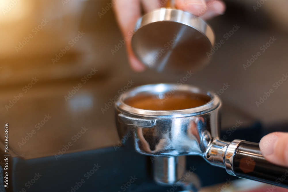 Barista woman is preparing, making espresso americano using professional machine, special tool kit in cafe. Closeup female hands are tempering, pressing ground coffee in metal portafilter.