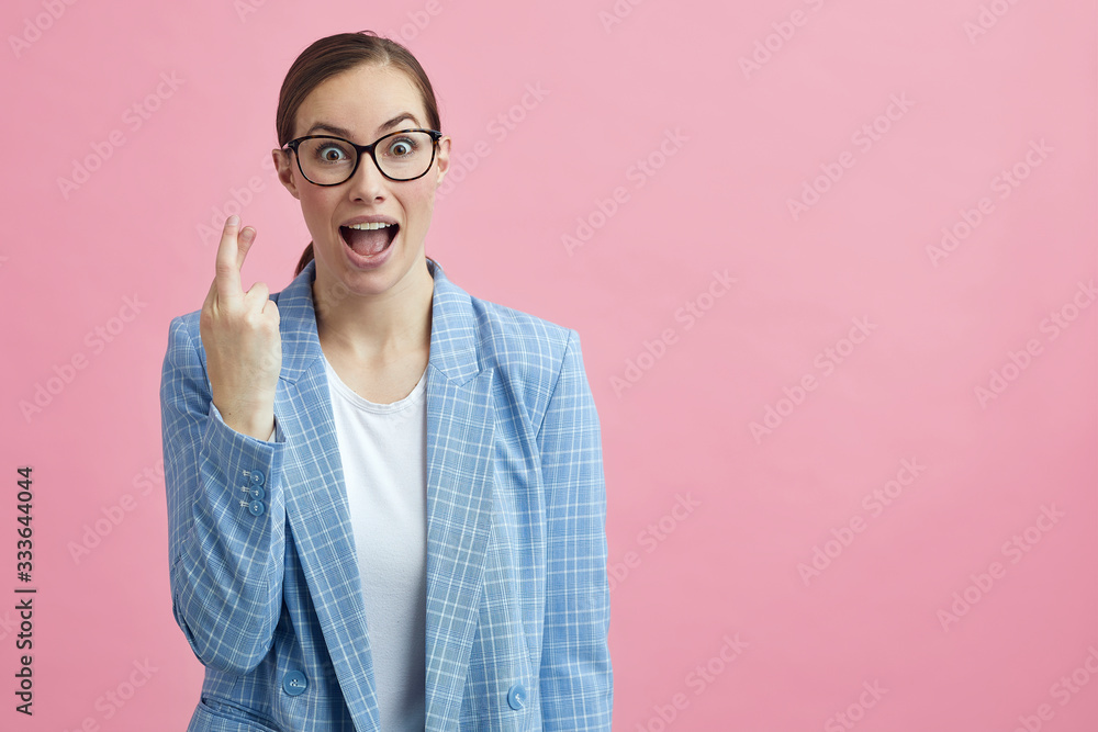 Businesswoman with glasses on a pink background, is excited and crossing her fingers hoping 