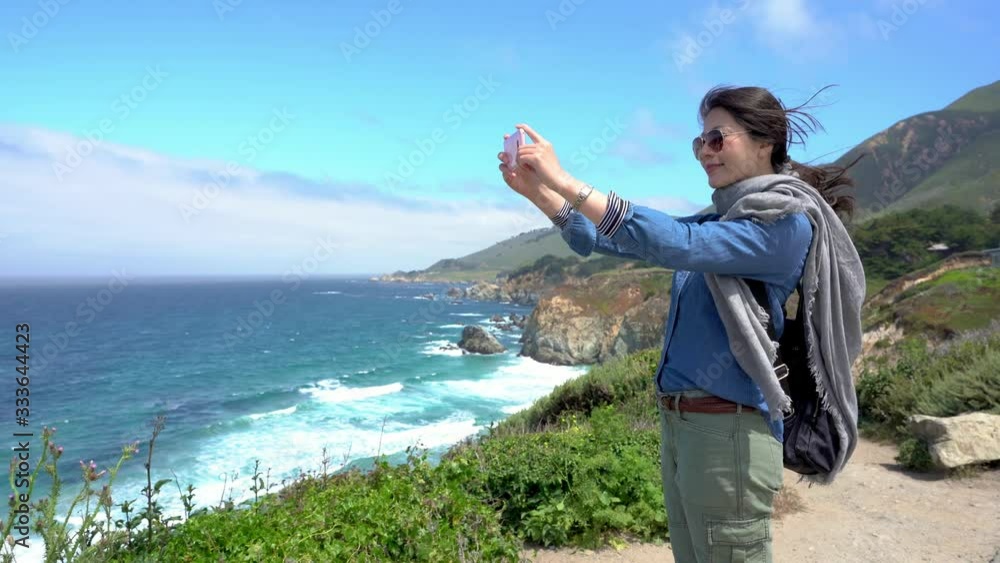 asian girl backpacker taking pictures of amazing landscape on mobile phone while standing on rock near sea. beautiful sunshine with sea and blue sky. Pacific coast highway 1 Big Sur California asian girl backpacker taking pictures of amazing landscape on mobile phone while standing on rock near sea. beautiful sunshine with sea and blue sky. Pacific coast highway 1 Big Sur California