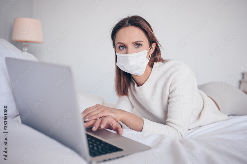 Young european woman in face medicine mask working on a laptop in bedroom during coronavirus isolation home quarantine. Covid-19 pandemic Corona virus. Distance online work from home concept.