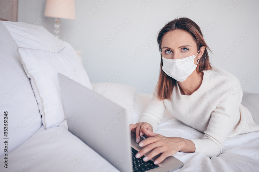 Young european woman in face medicine mask working on a laptop in bedroom during coronavirus isolation home quarantine. Covid-19 pandemic Corona virus. Distance online work from home concept. Young european woman in face medicine mask working on a laptop in bedroom during coronavirus isolation home quarantine. Covid-19 pandemic Corona virus. Distance online work from home concept.