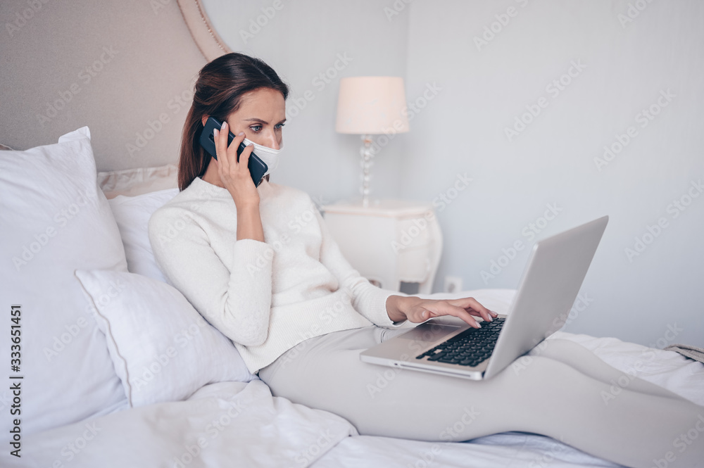 Young woman in bed, speaking phone and working on a laptop. Home quarantine self isolation during pandemic Corona virus. Distance work from home. COVID-19 concept to promote stay safe home save lives Young woman in bed, speaking phone and working on a laptop. Home quarantine self isolation during pandemic Corona virus. Distance work from home. COVID-19 concept to promote stay safe home save lives