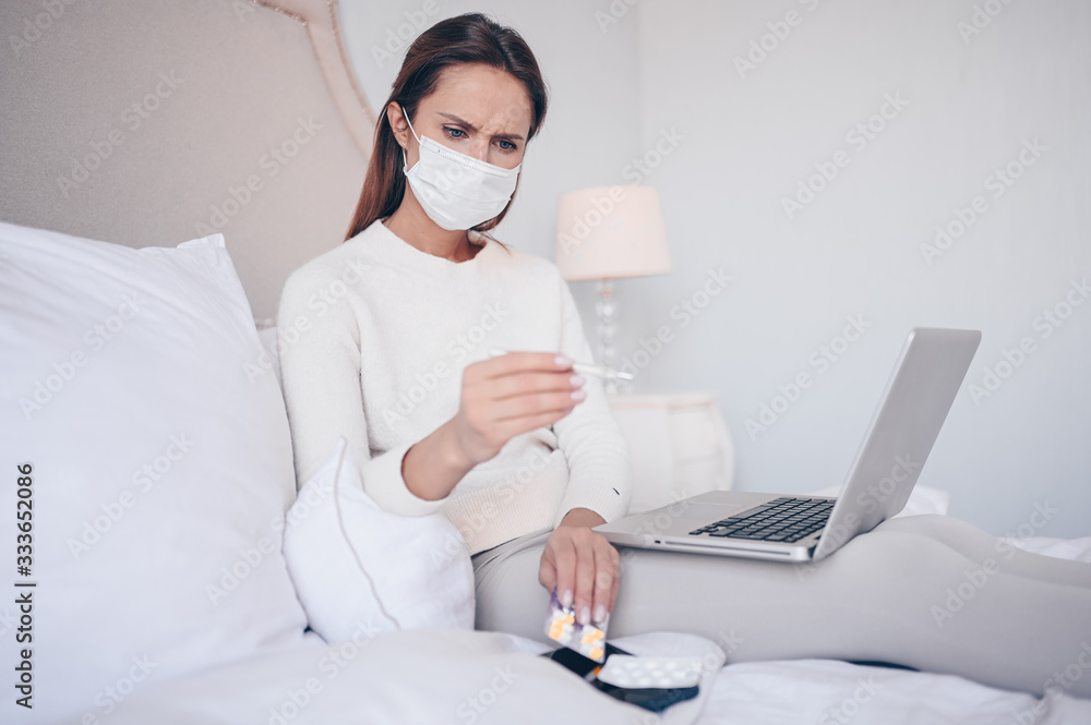 Sick woman in face protection mask lying in bed with laptop holding thermometer and pills at home quarantine isolation. Distance online work from home. Corona virus infection COVID-19 concept. Sick woman in face protection mask lying in bed with laptop holding thermometer and pills at home quarantine isolation. Distance online work from home. Corona virus infection COVID-19 concept.