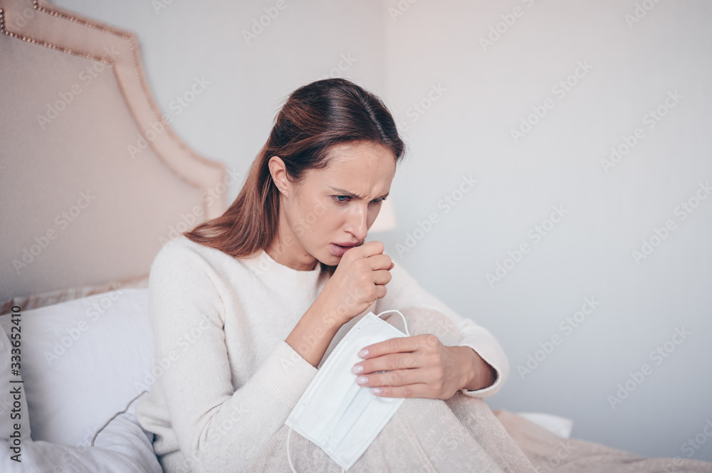 Coronavirus disease (COVID-19) symptoms - runny nose, sore throat, cough, fever. Young woman with face mask sick of flu viral infection spreading corona virus lying in bed at home quarantine isolation