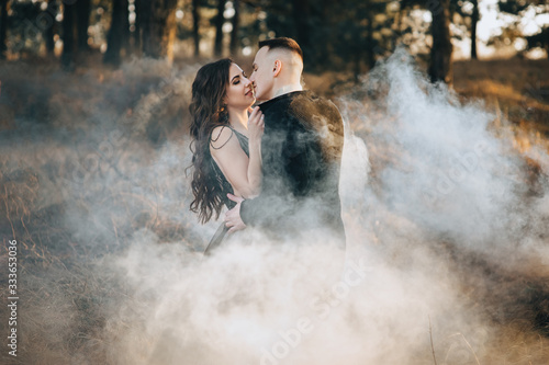A loving bridegroom in a black cardigan and a cute bride in an expensive dress are holding a pyrotechnic device with smoke in their hands and hugging. Wedding portrait of the newlyweds.