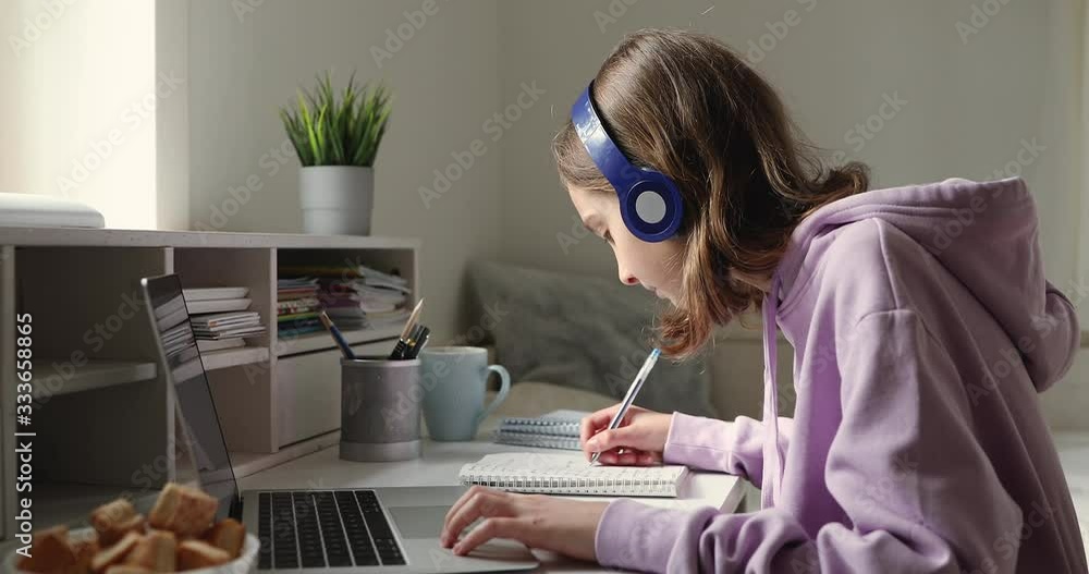 Teenage girl wearing headphones studying online from home with distance teacher. Teen school student distance learning in internet making notes, doing homework, watching video tutorial course concept.