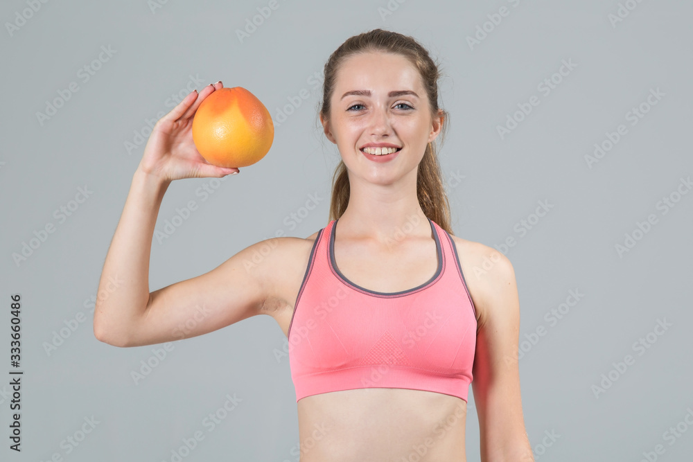 Young fitness woman holding a grapefruit in her hand, standing on a gray background and looking at the camera. Young fitness woman holding a grapefruit in her hand, standing on a gray background and looking at the camera.