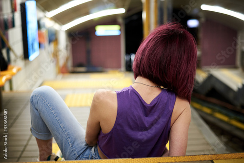 Young woman with burgundy pink maroon short hair, wearing purple top, light blue jeans and black sunglasses, sitting on stairs in subway station, Creative portrait of pretty girl in underground.