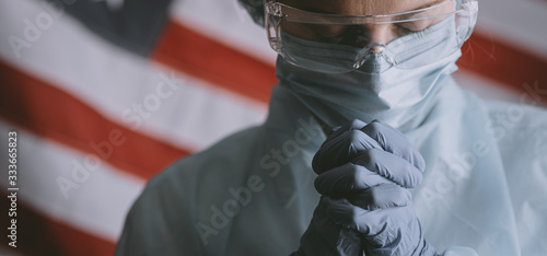 young nurse in a protective medical mask and hat and dressing gown prays against the backdrop of the American flag and asks God for help and strength in the fight against the coronavirus epidemic