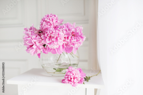 Pink peonies in glass vase. Flowers on white table near the window. Morning light in the room. Beautiful peony flower for catalog or online store. Floral shop and delivery concept. Banner. Copy space