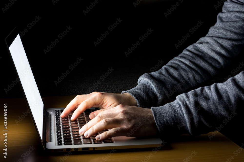 Man working on laptop with white screen glowing in the dark on a wooden desk from the side Man working on laptop with white screen glowing in the dark on a wooden desk from the side
