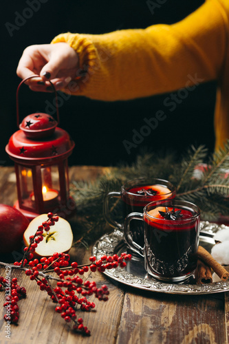 Mulled wine, fur-tree branches, red berries, woman's hand holding a candle. Cozy winter evening.