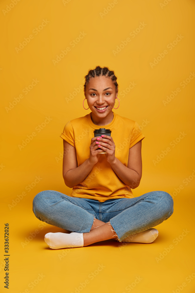 Relaxed dark skinned millennial girl sits in lotus pose, holds takeaway coffee, has casual talk with someone, wears yellow t shirt and jeans, smiles happily, enjoys spare time. People, lifestyle Relaxed dark skinned millennial girl sits in lotus pose, holds takeaway coffee, has casual talk with someone, wears yellow t shirt and jeans, smiles happily, enjoys spare time. People, lifestyle