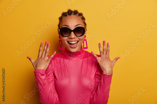 Portrait of happy positive dark skinned woman raises hands, shows perfect rosy manicure, wears sunglasses and fashionable outfit, smiles positively, isolated on yellow background, enjoys sunny day