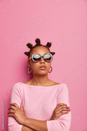 Self confident dark skinned woman keeps arms folded, stands in assertive pose, has many knots on head, wears sunglasses and rosy jumper, thinks about something, poses indoor. People, style, vogue