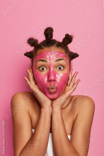 Fototapeta Naklejka Na Ścianę i Meble -  Portrait of beautiful female touches cheeks gently, keeps lips folded, applies beauty mask on face to rejuvenate and tone skin, isolated on pink background. People, relaxation, cosmetic treatment